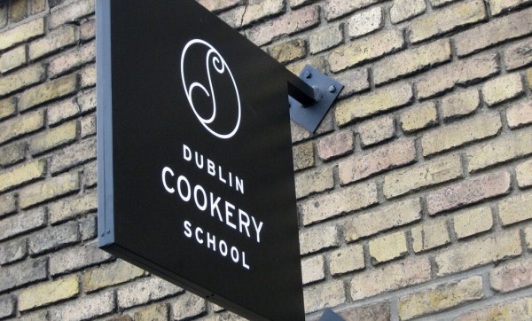 A black projecting sign that displays the Dublin Cookery School Logo outside their building